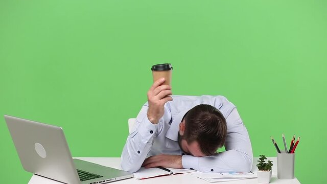 Young weary exhausted tired employee man sit work laid his head down sleep on hands hold up cup of coffee tea at desk pc laptop computer isolated on green background Business career lifestyle concept