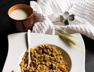 Cereal granola in a white plate with a spoon and a clay glass of milk on a black wooden tabletop.
