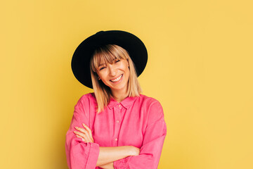 Cheerful positive beautiful young woman posing on camera alone in studio. Girl smile and hld hands crossed. Model in pink shirt and black hat isolated over yellow background.