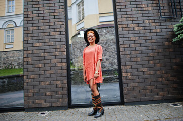 African red haired woman wear eyeglasses, orange dress and black hat posing outdoor.