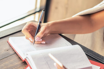 Hand of business woman writing in diary by pen using smartphone near window in the cafe.