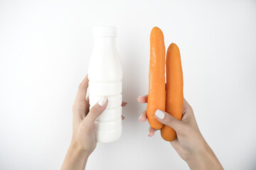image of woman's hands holding yogurt in plastic bottle in one hand and two fresh carrots in another on isolated white background