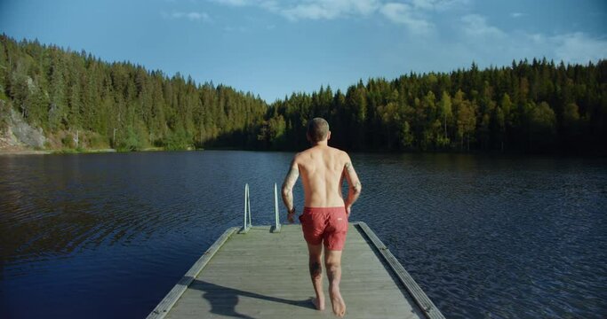 Millennial young man with tattoos run on wooden dock or pier and jump into refreshing cool water of mountain forest lake. Concept staycation adventures and summer time activities in backyard 