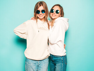 Two young beautiful blond smiling hipster girls in trendy summer hoodie clothes. Sexy carefree women posing near blue wall. Trendy and positive models having fun in sunglasses