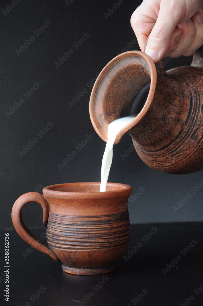 Wall mural Milk flowing from a clay jug in a man’s hand onto a clay mug on a black background