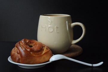 Saucer with a bun and a teaspoon and a mug with a drink in defocus in the dark background