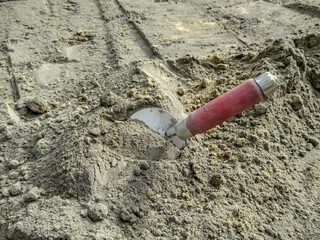 Steel trowel with a red wooden handle stuck in a gray dry mortar. Hand tool for mixing sand and cement for construction work