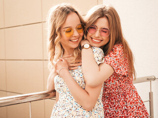Two young beautiful smiling hipster girls in trendy summer sundress.Sexy carefree women posing on street background in sunglasses. Positive models having fun and hugging
