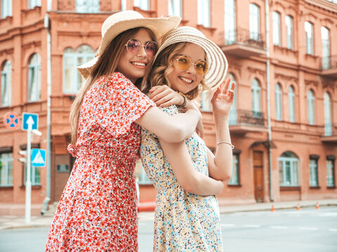 Two young beautiful smiling hipster girls in trendy summer sundress.Sexy carefree women posing on street background in hats. Positive models having fun and hugging