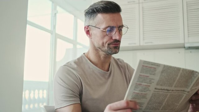 A serious man is reading a newspaper while eating breakfast at home in the kitchen at the morning