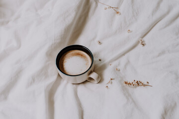 Obraz na płótnie Canvas Cozy black and white cup of coffee between bed sheets decorated with some dried flowers. Relaxing photo. Morning coffee concept.