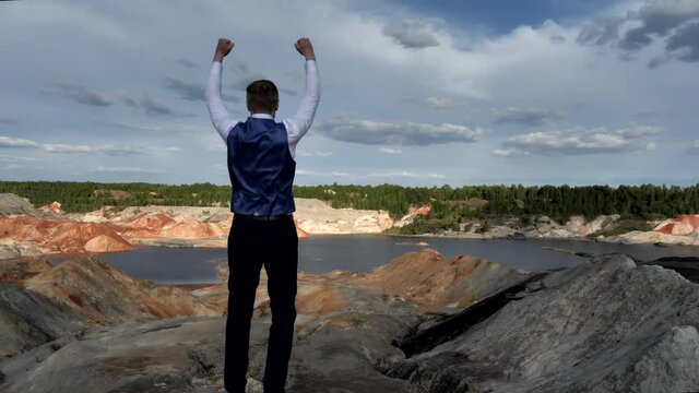 The camera approaches a businessman who stands on the edge of a hill and triumphantly throws up his hands looking at the wide expanse of a hilly Martian landscape with a small pond, the camera stops