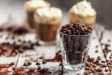 Coffee beans in a glass cup and in the background a cup with coffee and whipped cream