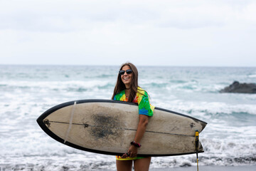 Surf girl in a flashy shirt holding a surfboard on the beach