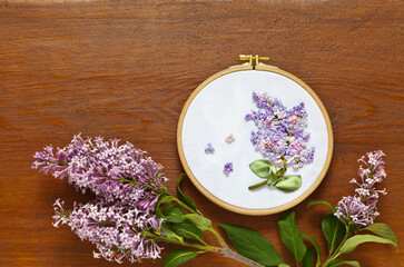 Obraz na płótnie Canvas Needlework and hobby. Top view on embroidery with satin ribbons of a sprig of blooming lilac in a round frame on a wooden background. Handmade gift. Flat lay, copy space, close-up, mock up
