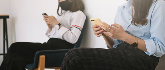 Fototapeta premium Two Asian young woman chatting in smartphone and wearing mask sitting distance protect from COVID-19 viruses for social distancing for infection risk.