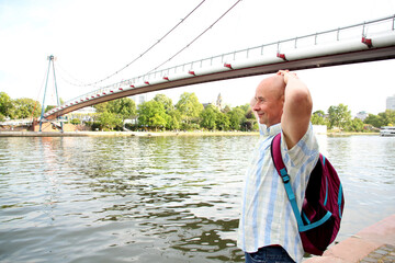 old satisfied man, tourist with a backpack, stands at the bridge on the banks of the Main river in...