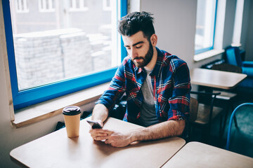 Thoughtful hipster guy looking at cafeteria window while waiting for call from friend on new...