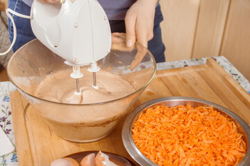 Kobieta przygotowuje ciasto marchewkowe. Dłoń kobiety trzyma mikser i miksuje składniki. Starta marchewka leży na misce. Skorupki jajek leżą na talerzyku.