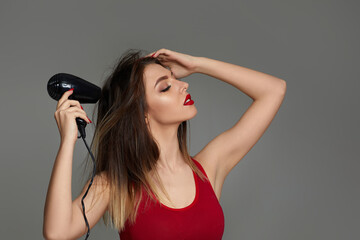 Beautiful young woman drying her hair with dryer on gray background