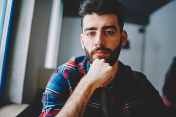 Portrait of concentrated handsome hipster guy 20 years old sitting indoors and looking in camera...