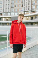 Obraz premium Street portrait of a sporty young man in a red hoodie and shorts standing against the backdrop of a velodrome and a building and looking at the camera with a serious face. Athletic guy posing.