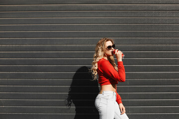 Fototapeta premium A young woman with a perfect body in a red sweatshirt is drinking cool soft drink on a summer sunny day outdoors.