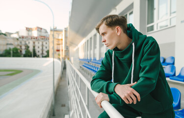 Athletic young man in a green sports suit stands in the stands and looks away at the velodrome with a sporty face. Athletic guy rests after training. Copy space