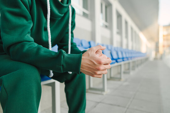 Close-up, cropped photo. Athletic man in a green tracksuit sitting in the stands and resting after training, focus on his hands. Background.