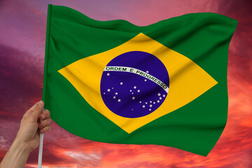 male hand holds Brazil national flag on sky background with clouds on luxury satin texture, silk with waves, closeup, copy space, concept of travel, economy, politics