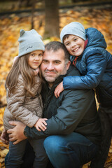 A boy and a girl hug a man, their dad, they are smiling. In the background autumn forest. They are warmly dressed in jackets and hats. Soft focus.
