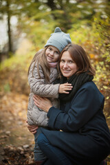 Mom and daughter hug each other, mom crouched. A woman in a black coat, a girl in a jacket and hat. In the background is golden autumn. Soft focus.