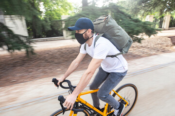 Obraz premium Repartidor de comida a domicilio con mascarilla en bicicleta amarilla por la ciudad con la mochila cargada de pedidos