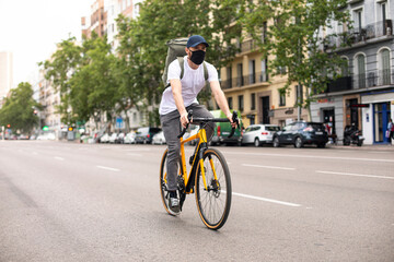 Repartidor de comida a domicilio con mascarilla en bicicleta amarilla por la ciudad con la mochila cargada de pedidos