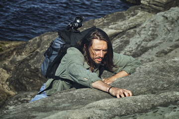 Naklejka na ściany i meble young long-haired guy with a backpack and a photo tripod climbs on a steep rock on the seashore. Climbing and outdoor concept.