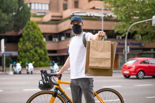 Repartidor de comida a domicilio con mascarilla en bicicleta amarilla por la ciudad con la mochila cargada de pedidos mostrando bolsas de cart&oacute;n