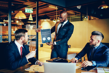 Confident intelligent male entrepreneur holding infographic with data and accountings speaking during meeting with colleagues, dark skinned financial expert analyzing corporation income with partners