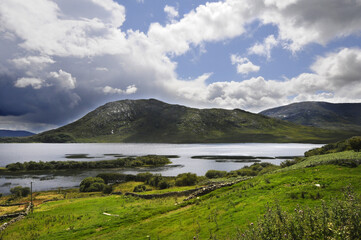 Vue panoramique sur un lac et ses îlots de terre, avec une ruine de tour médiévale en son centre. Ciel chargé et nuageux, collines, verdure et rochers.