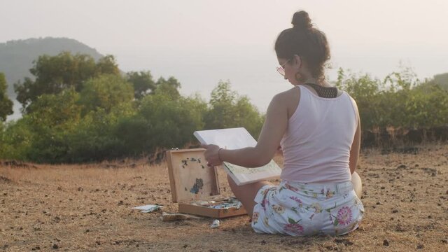 Back view of young artist painting picture outdoors in mountains background slow motion. Woman sitting with canvas and art tools working alone on nature. Creative process meditation relaxation