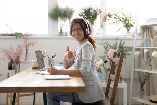 Portrait of smiling young woman in headset sit at desk studying show thumb up recommend online course, happy millennial female give recommendation to distant training or class, education concept