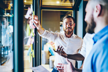 Cheerful bearded proud ceo in white shirt laughing while pointing on glass wall with colorful...
