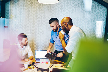 Multiethnic business men drinking coffee and discussing graphs in office with laptop on table