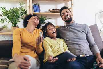 Fototapeta premium Cute little Afro-American girl and her beautiful young parents using digital tablet and doing shopping online while sitting on a sofa at home.