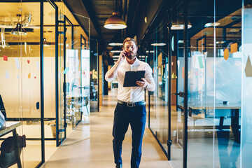 Busy bearded office employee with black folder in hand calling on smartphone going on office corridor of finance company.Caucasian male proud ceo dressed in formal wear talking on cellular