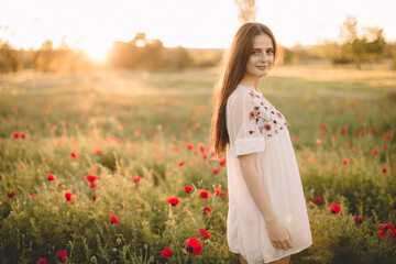 Obraz na płótnie Canvas Young happy smile caucasian girl staying at a green red poppy field in the morning. The background of the sun shines. Concept of freedom. Summer. Beautiful young woman walking on field