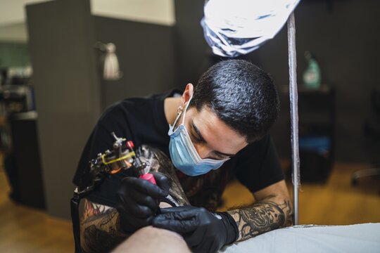 Shot Of A Male Tattoo Artist Wearing A Mask And Gloves And Tattooing His Client's Leg