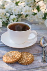 White porcelain cup with tea and two oatmeal cookies with sesame seeds on a wooden table against the background of blooming jasmine.