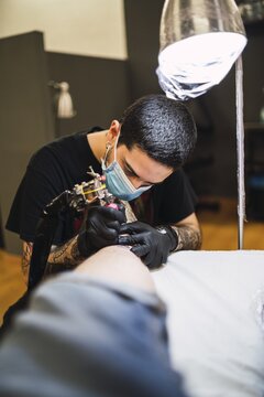 Shot Of A Male Tattoo Artist Wearing A Mask And Gloves And Tattooing His Client's Leg