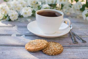 White porcelain cup with tea and two oatmeal cookies with sesame seeds on a wooden table against the background of blooming jasmine.