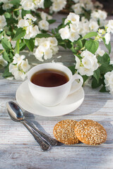 White porcelain cup with tea and two oatmeal cookies with sesame seeds on a wooden table against the background of blooming jasmine.